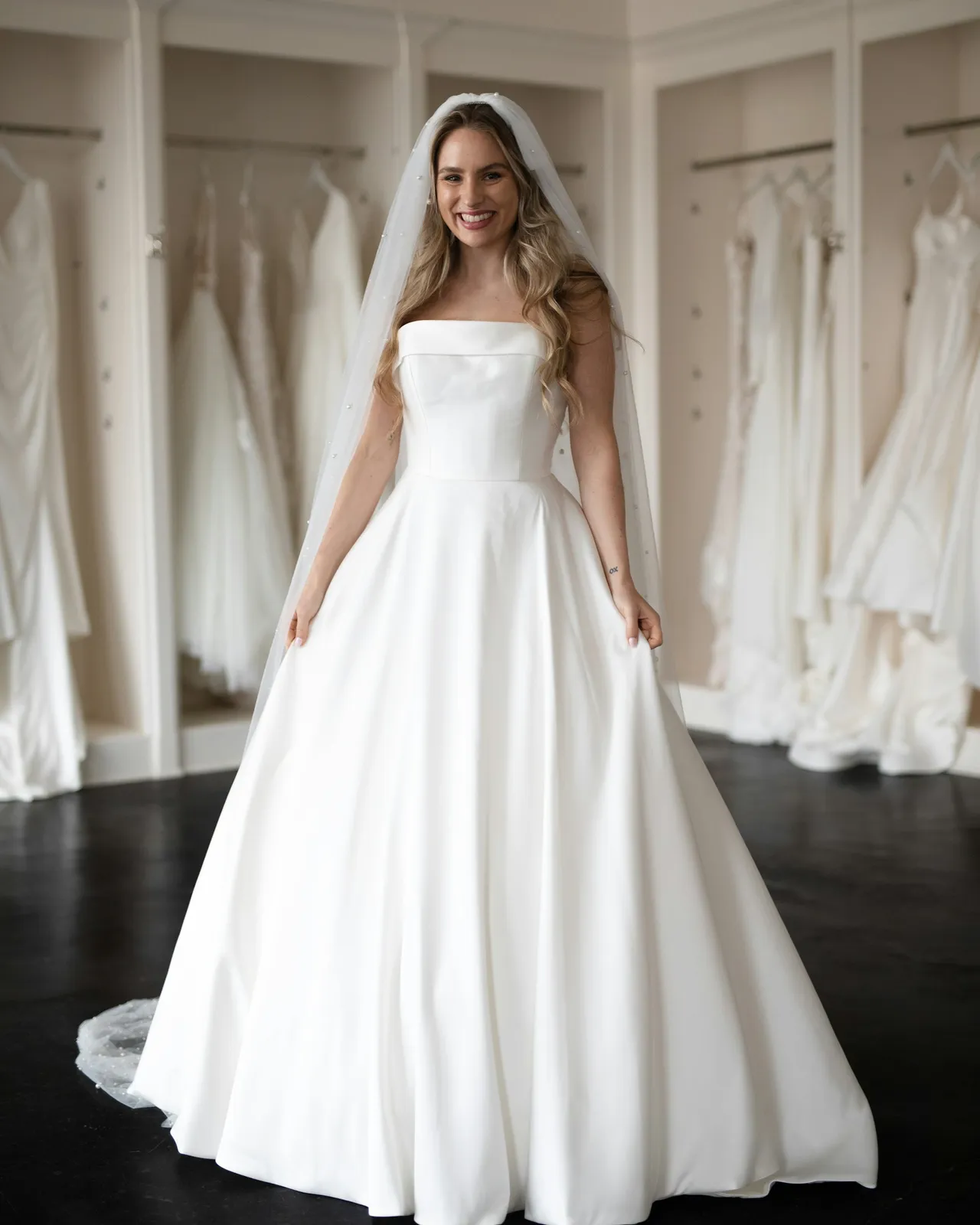 A bride smiling in a strapless satin ball gown in a bridal salon